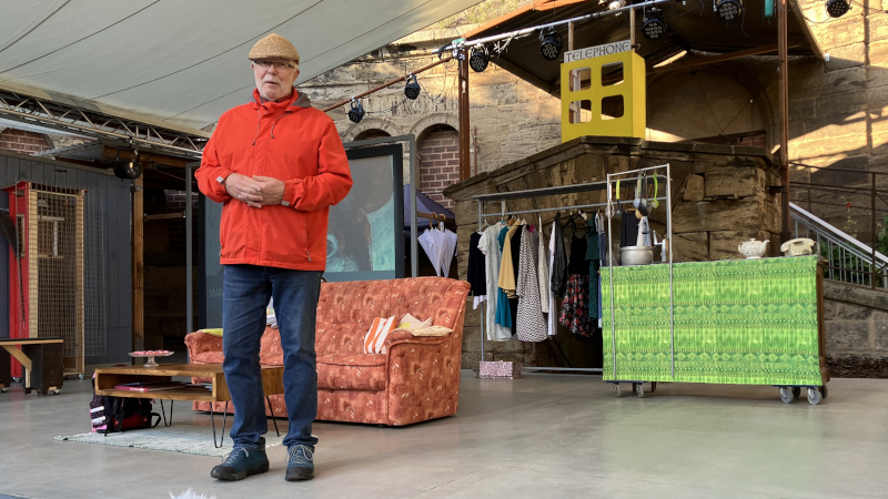 Bühnenbild mit rotem Sofa, kleinem Holztisch und Kleiderständer vor einer Kulisse aus Stein mit beleuchtetem „TELEPHONE“-Schild; im Vordergrund steht eine Person in roter Jacke und Jeans. Die Szene wirkt wie eine inszenierte Theater- oder Kulturveranstaltung.